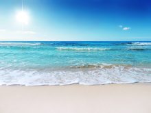 The sun is beating down on a white sand beach with the blue and green ocean in the background.