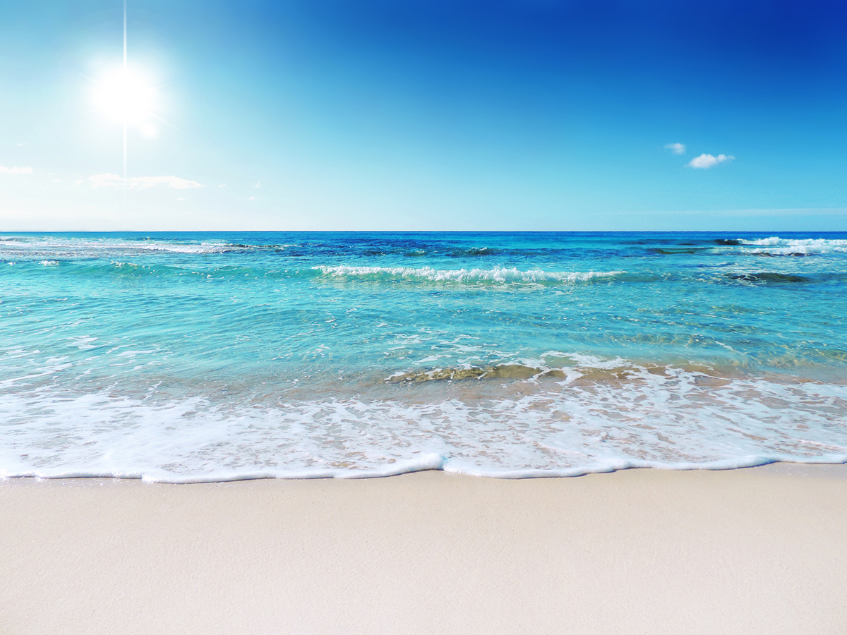 The sun is beating down on a white sand beach with the blue and green ocean in the background.