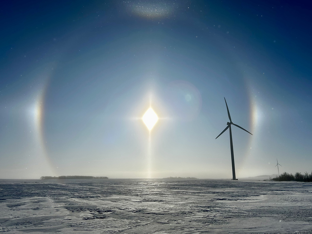 A perfect circle is scribed in the crisp blue sky by the sun and its sun dogs with a giant wind turbine in the foreground on a bitterly cold day.