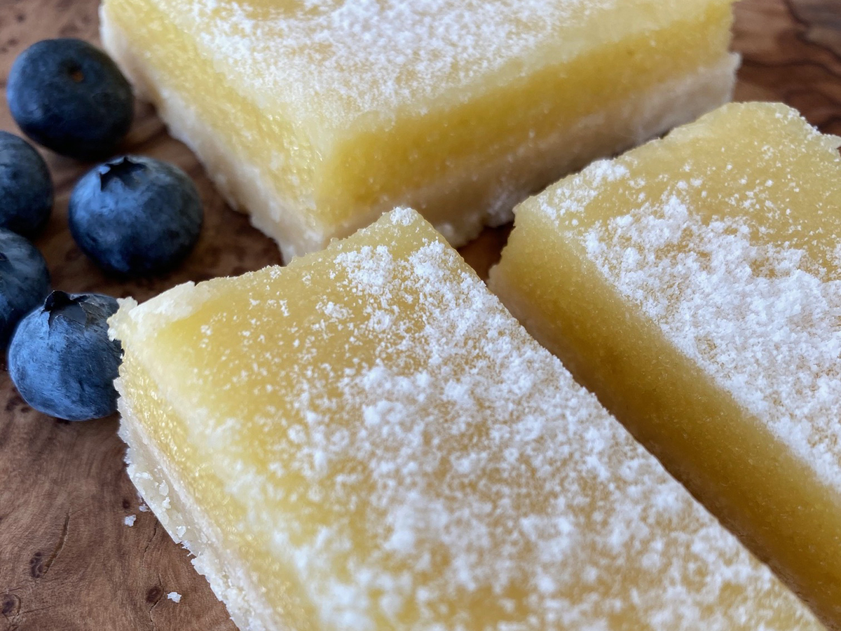 A close-up of three lemon squares dusted with icing sugar on a wooden surface with five juicy blueberries beside them.