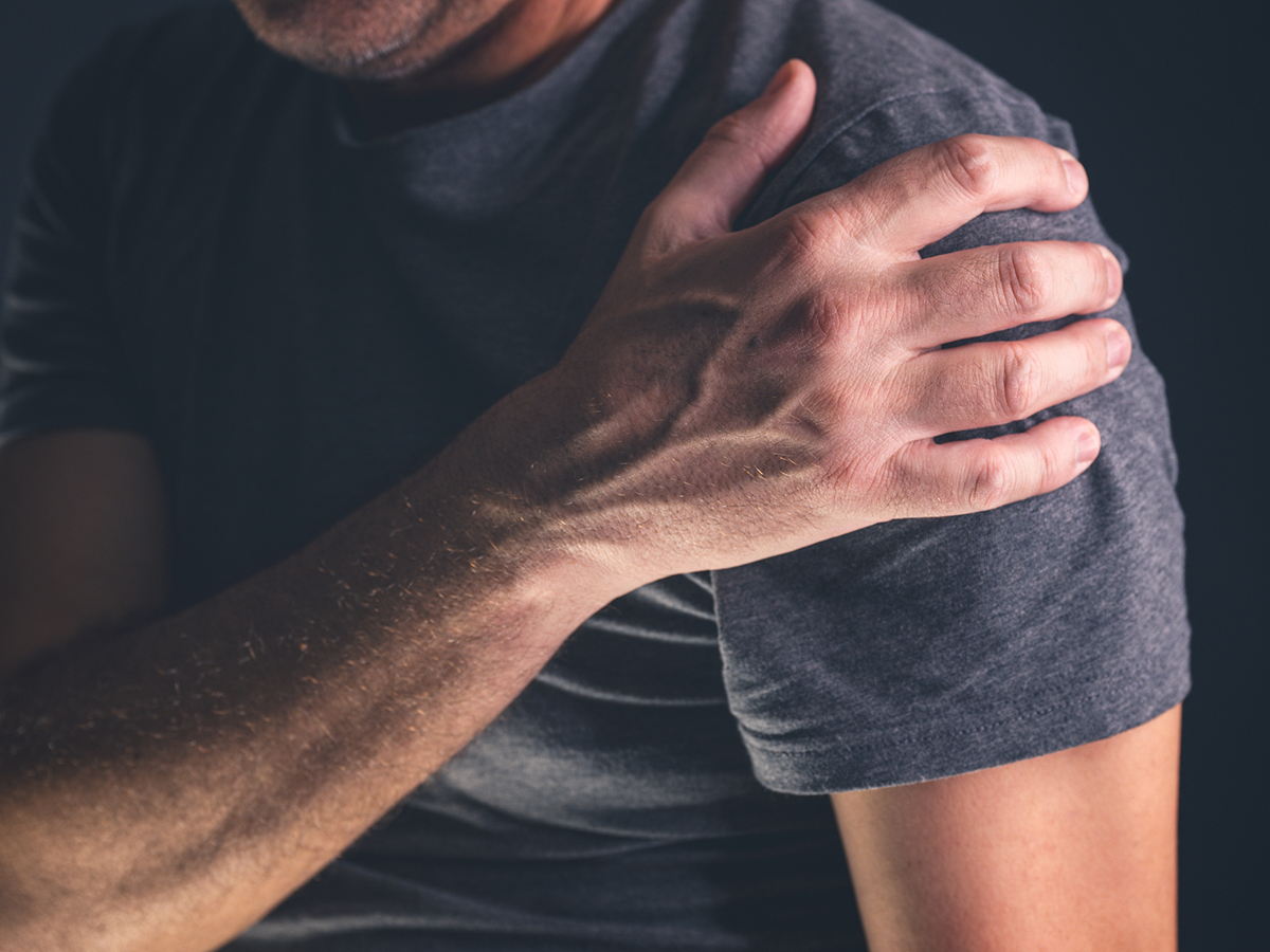 A close-up of a t-shirt-wearing man's right hand grabbing his left shoulder.