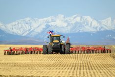 A large tractor pulls a massive air seeder rig directly toward the camera seeding into stubble with the snow-capped Rocky Mountains in the background.